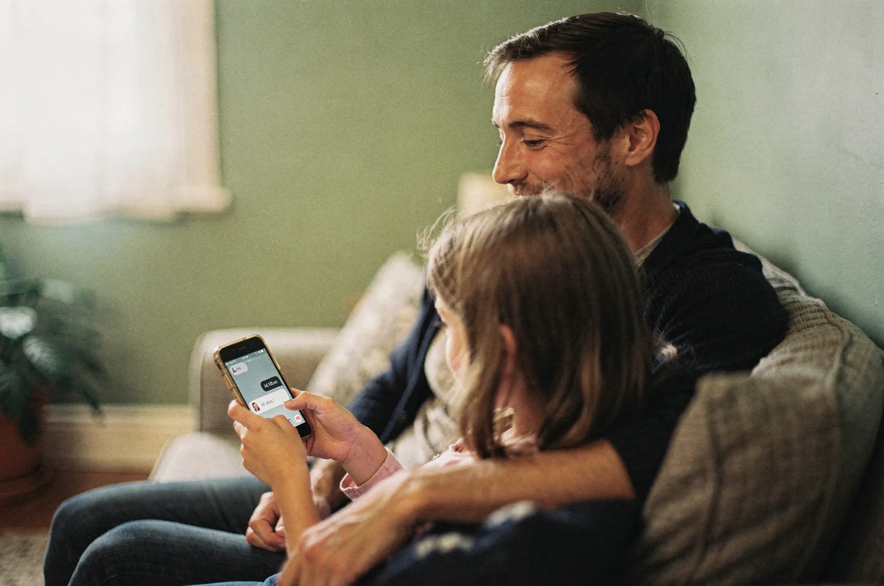 A parent and a primary-school-age child sitting close on a sofa, sharing an older phone the child is holding.