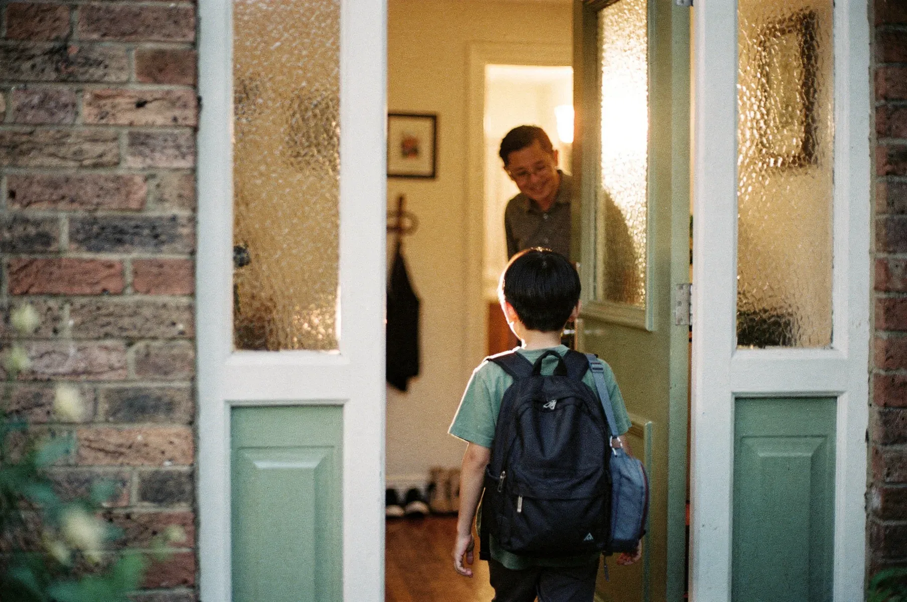 A child with a backpack, seen from behind, stands at an open front door as a parent welcomes them in from a warmly lit hallway.