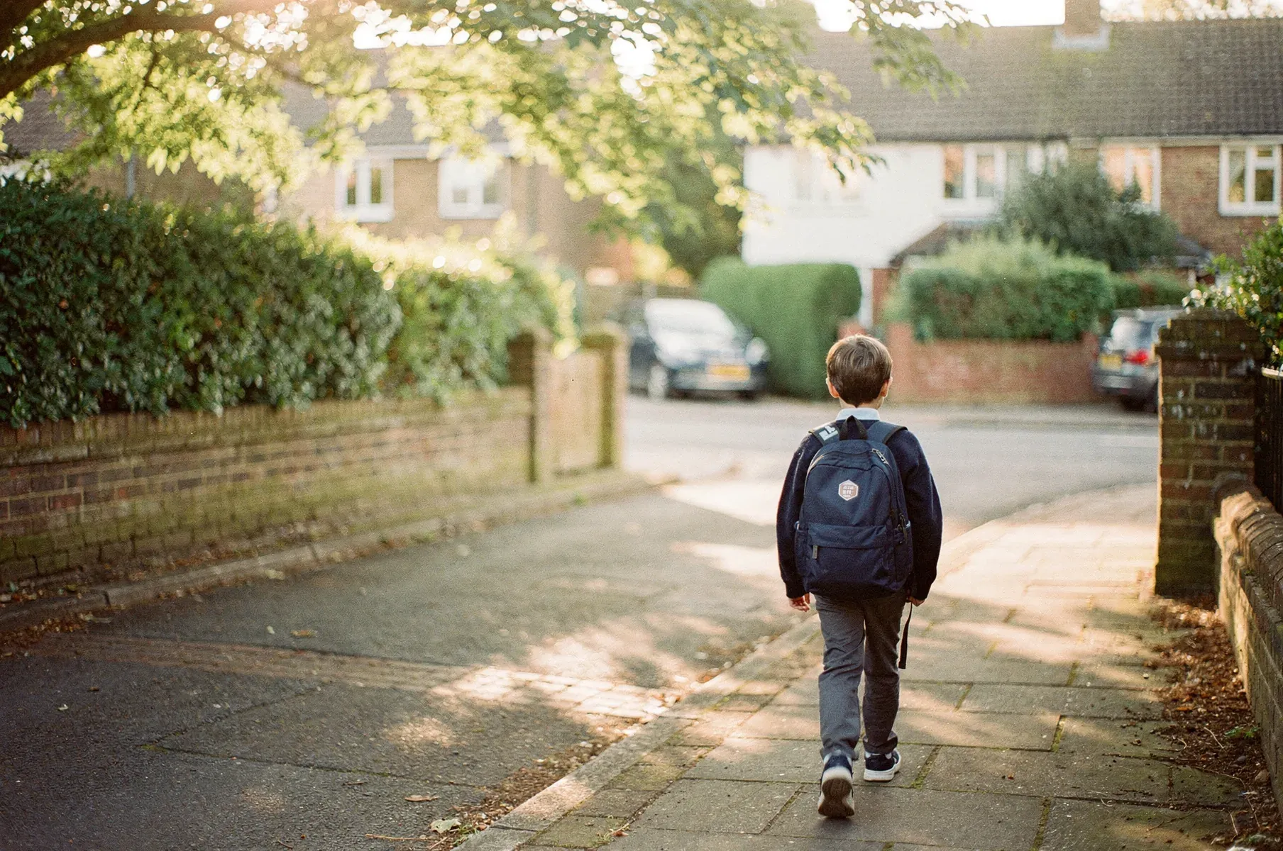 A primary-school-age child walks away from the camera down a quiet, sunlit suburban street, seen from behind with a navy school backpack.