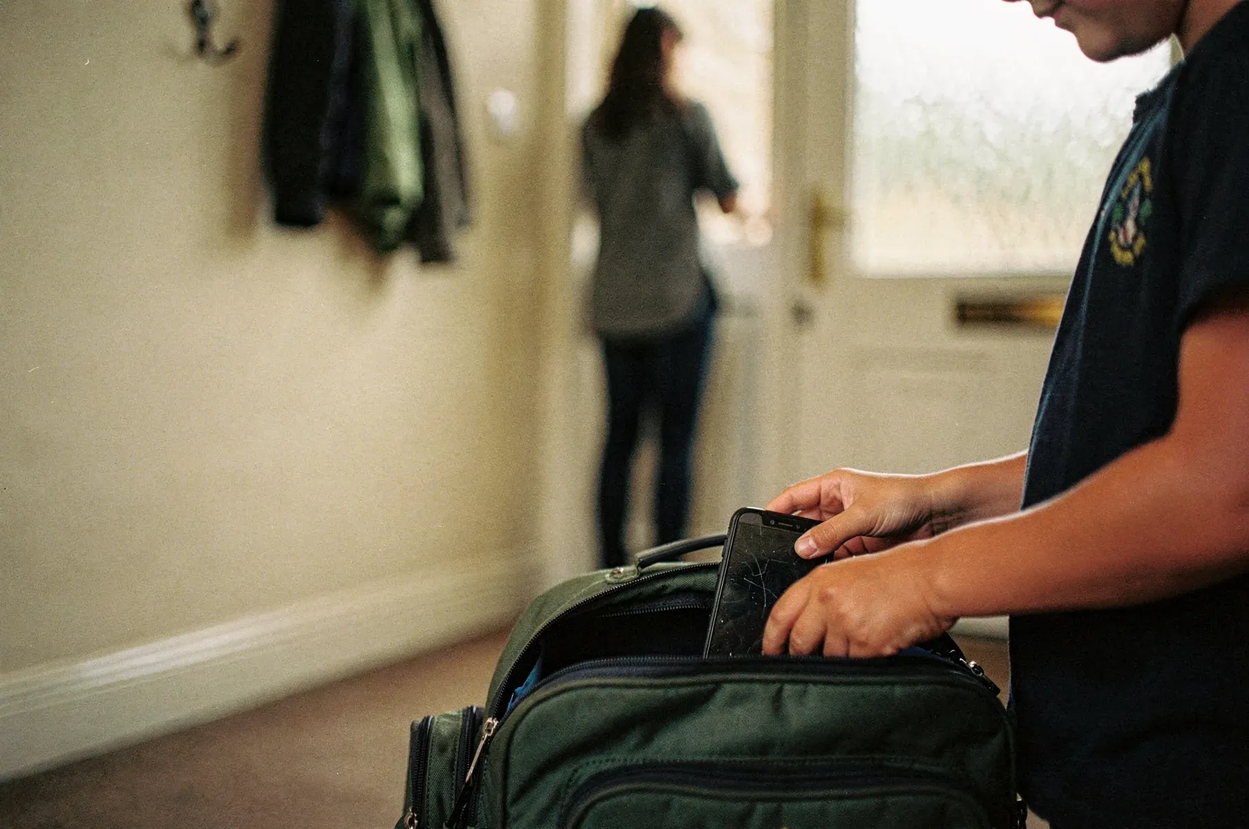 A child slides an older phone into a green school backpack by the front door, their face out of frame, a parent softly blurred in the hallway behind.