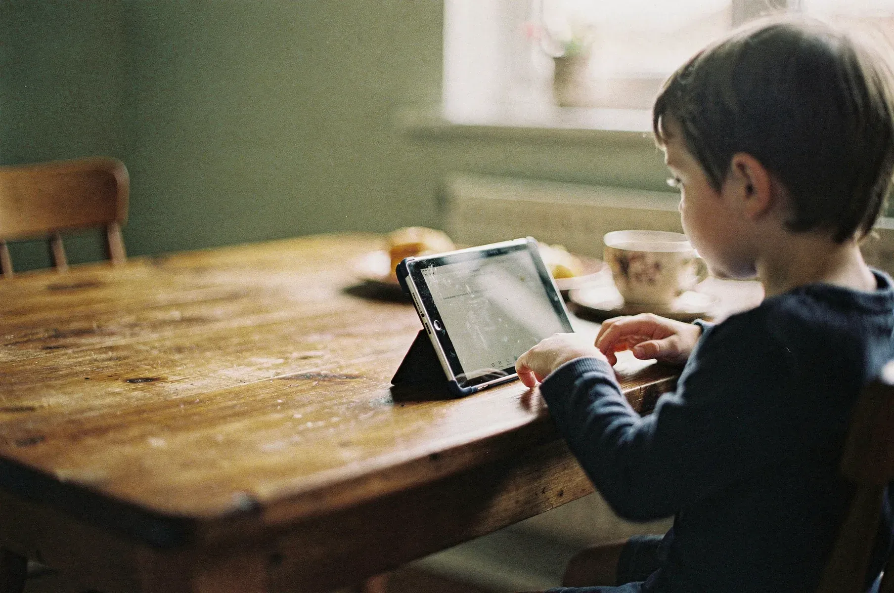 A young child sits at a wooden kitchen table in soft morning light, looking at a tablet propped up in front of them beside a cup and breakfast things.